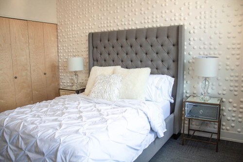 A cozy bedroom featuring a tufted gray headboard, white bedding, and stylish lamps beside a mirrored nightstand.