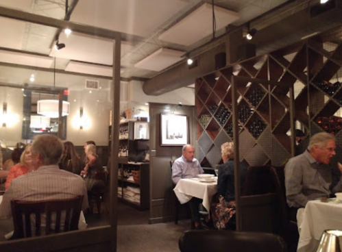 A cozy restaurant interior with diners at tables, soft lighting, and a wine display in the background.