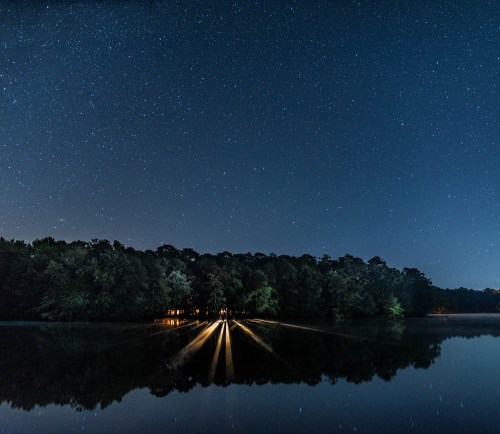 A serene night scene with a starry sky reflecting on a calm lake, surrounded by trees and soft lights along the shore.