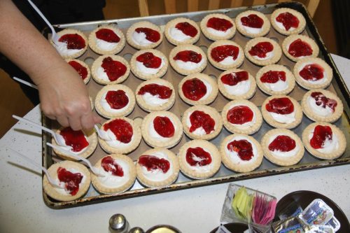 A tray of mini dessert cups filled with whipped cream and topped with red jelly, being arranged by a hand.