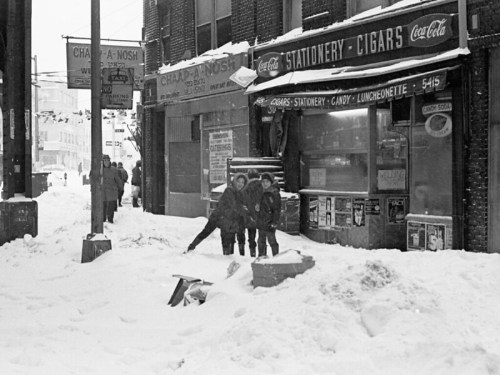Three children play in deep snow outside a storefront, with snow-covered streets and buildings in the background.
