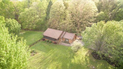 Aerial view of a cozy brown house surrounded by lush green trees and a spacious lawn.