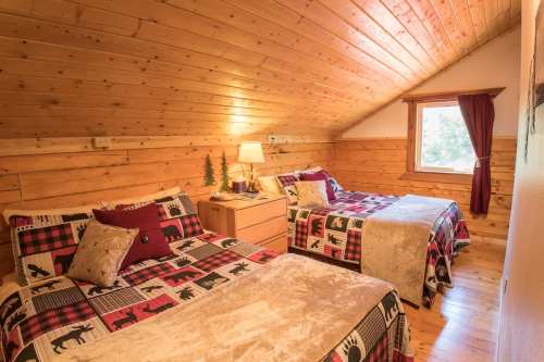 Cozy attic bedroom with two beds, wooden walls, and warm lighting, featuring rustic decor and a window.