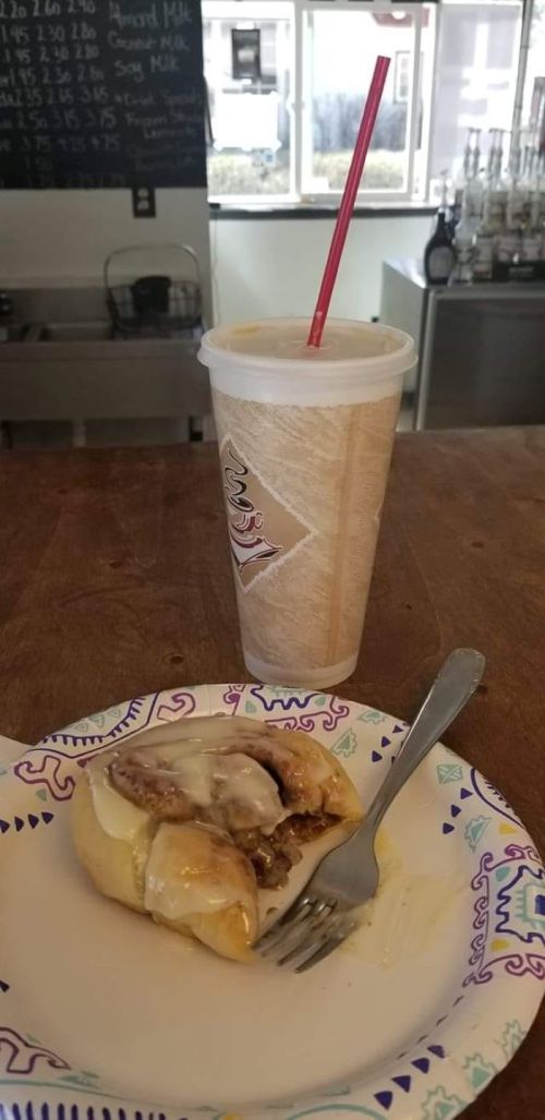 A plate with a cinnamon roll and a fork beside a large drink with a straw on a wooden table.