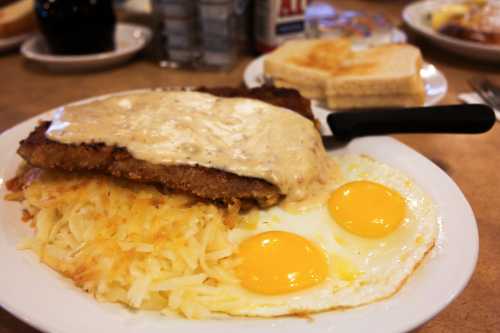 A plate of breakfast featuring eggs, hash browns, toast, and a breaded meat patty topped with creamy gravy.