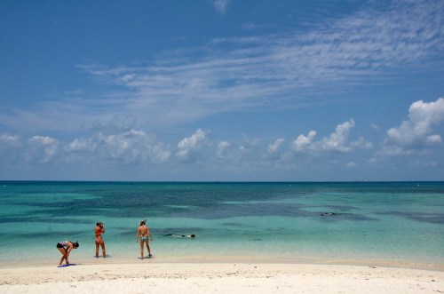 Three people on a sandy beach, with clear turquoise water and a blue sky dotted with clouds in the background.