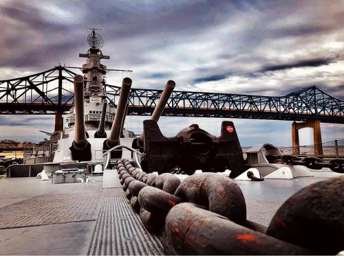 A battleship's bow with large cannons and an anchor chain, set against a cloudy sky and a bridge in the background.