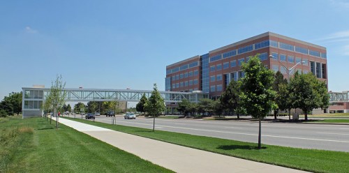 A modern building with large windows, connected by a walkway, alongside a tree-lined street on a sunny day.