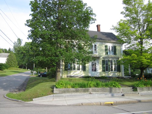 A green two-story house with large windows, surrounded by trees and a well-maintained lawn, on a quiet street.