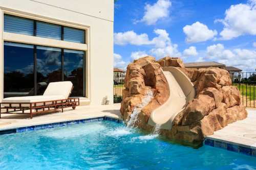 A pool with a rock-style waterslide, surrounded by a patio and a lounge chair, under a blue sky with clouds.