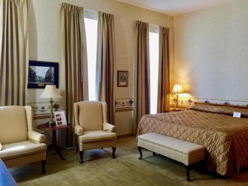 Cozy hotel room with a bed, two armchairs, a bench, and soft lighting from lamps by the windows.