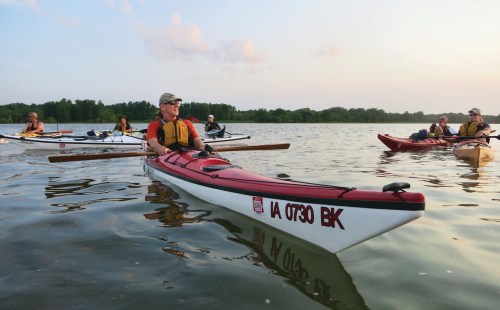 A group of kayakers paddles on calm water during sunset, surrounded by trees and a serene landscape.