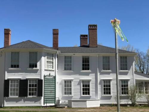 A white two-story house with multiple chimneys and a flagpole adorned with colorful ribbons against a clear blue sky.