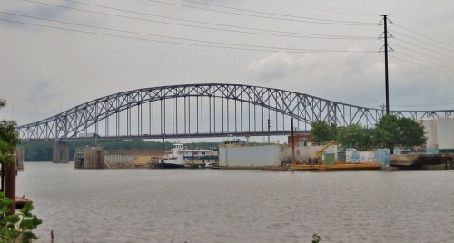 A large steel bridge spans a river, with boats docked and industrial buildings along the shore under a cloudy sky.