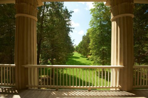 View from a porch with columns, overlooking a lush green lawn and trees under a bright blue sky.