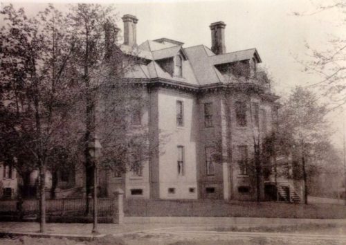 Historic black-and-white photo of a large, ornate building surrounded by trees and a fence.