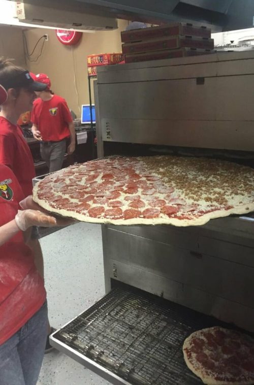A worker pulls a large pepperoni pizza from an oven, with another pizza on the rack below.