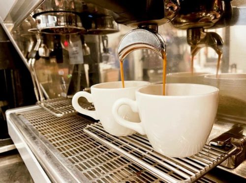 Two white coffee cups sit under an espresso machine, with rich coffee pouring into them.