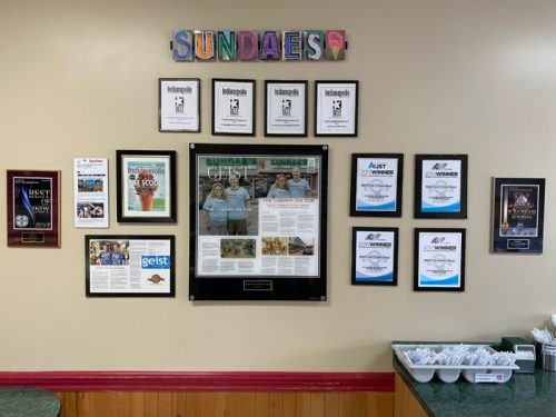 A wall display featuring awards, certificates, and a framed newspaper article in a colorful ice cream shop.