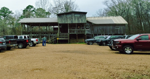 A gravel parking lot filled with trucks and SUVs in front of a large wooden building surrounded by trees.