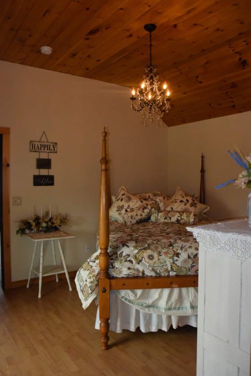 Cozy bedroom with a four-poster bed, floral bedding, a chandelier, and a small table with decorative flowers.