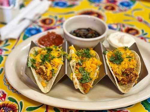 Three tacos filled with meat, cheese, and greens, served with black beans, salsa, and sour cream on a colorful table.