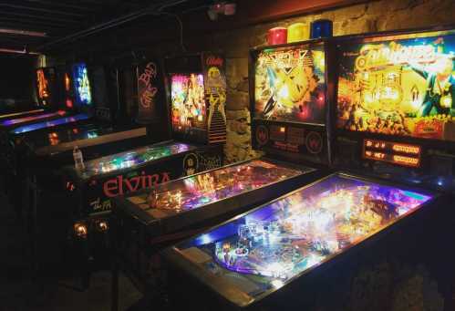 A dimly lit arcade featuring several colorful pinball machines with vibrant lights and retro designs.