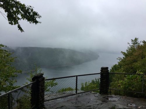 Foggy landscape view of a lake surrounded by lush greenery and hills, with a stone railing in the foreground.
