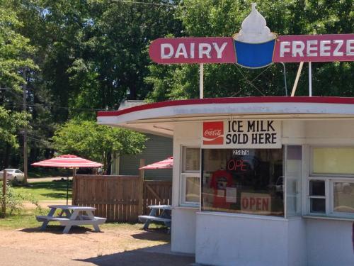 A quaint ice cream stand with a red and white exterior, featuring picnic tables and umbrellas in a green setting.