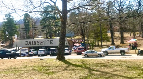 A view of a roadside diner named "Dairyette" with parked cars and trees in the background on a sunny day.