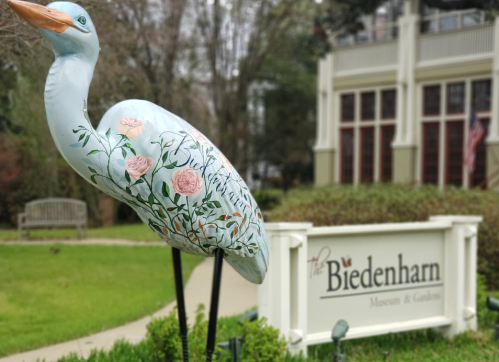 A painted heron statue with floral designs stands in front of the Biedenharn Museum and Gardens sign.