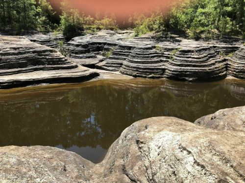 Rocky landscape with layered formations and a calm water pool surrounded by greenery.