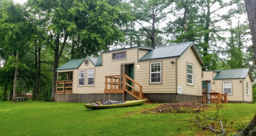 A cozy cabin with a green roof, surrounded by trees, and a kayak resting on the grass in front.