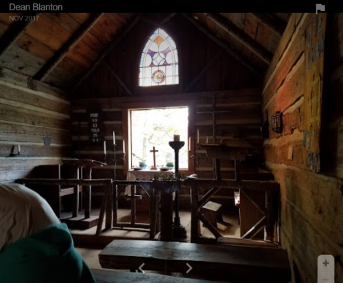 Interior of a rustic wooden cabin with a stained glass window, wooden benches, and a small altar. Natural light filters in.
