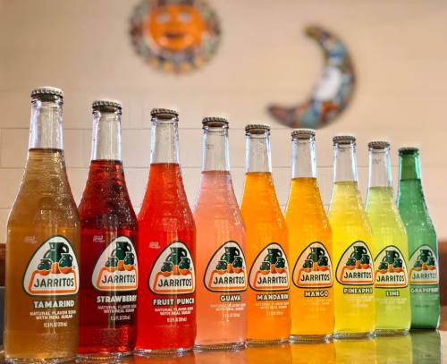 A colorful display of Jarritos soda bottles in various flavors, arranged in a row on a wooden surface.