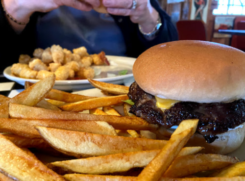 A close-up of a burger with fries in the foreground, and a plate of tater tots in the background.