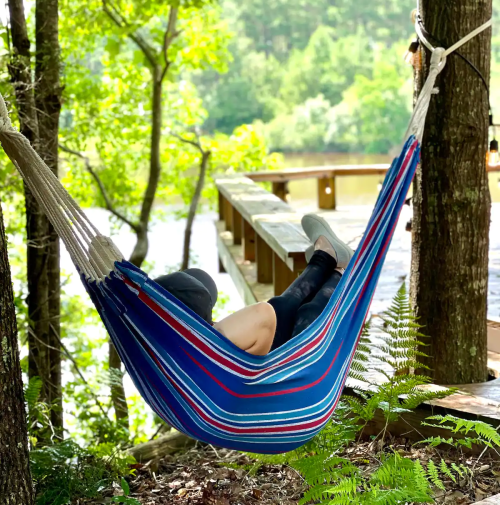 A person relaxes in a colorful hammock between trees, with a serene lake and wooden path in the background.