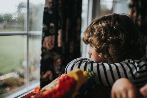 A child with curly hair gazes out a rainy window, resting their chin on their arms, lost in thought.