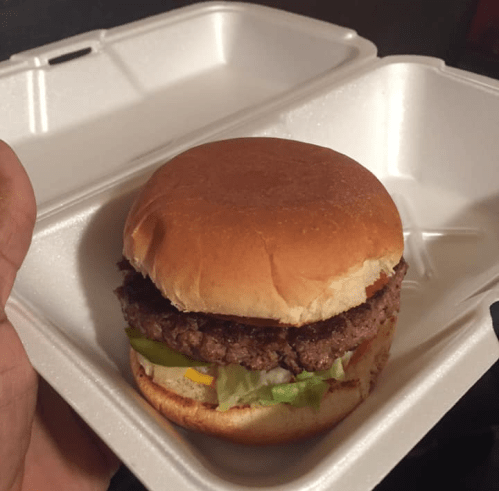 A hand holding a burger in a takeout container, featuring a beef patty, lettuce, and a soft bun.