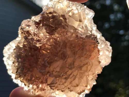 A hand holds a translucent, sparkling crystal cluster with shades of brown and gold, illuminated by sunlight.