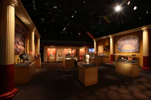 A museum exhibit featuring ancient artifacts displayed in a dimly lit room with columns and spotlighting.