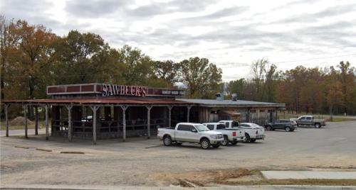 A rustic restaurant named "Sawbuck's" with a gravel parking lot and several parked trucks, surrounded by trees.