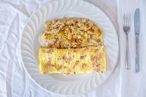 A plate with a yellow omelet filled with ham and a side of scrambled eggs, accompanied by a fork and knife.