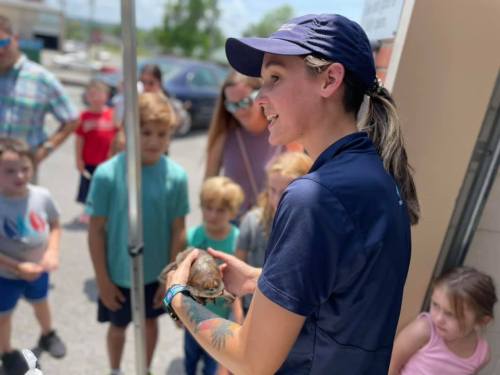 A woman in a cap holds a turtle while engaging with a group of children outdoors.