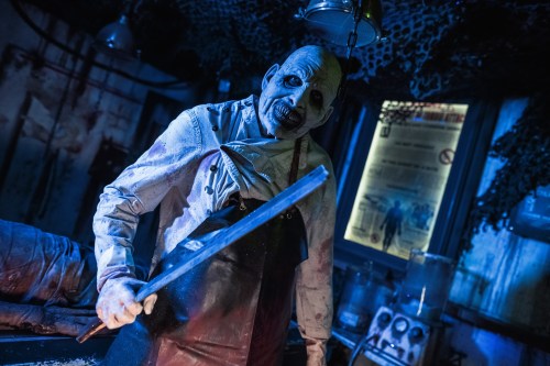 A creepy, lifelike figure in an apron holds a large knife in a dimly lit, eerie setting.