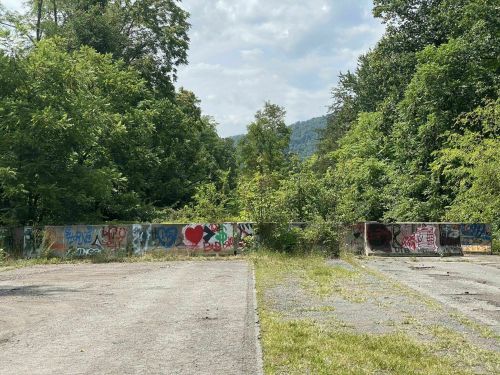A view of a grassy area with a graffiti-covered wall, surrounded by trees and mountains under a cloudy sky.