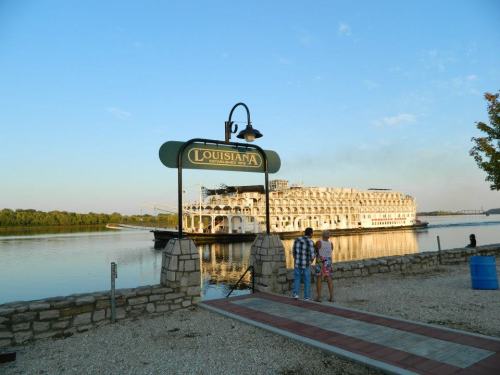 A couple stands by a sign reading "Louisiana" near a riverboat on calm waters, with trees in the background.