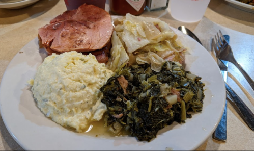 A plate of food featuring sliced ham, mashed potatoes, cabbage, and collard greens, with utensils on the side.