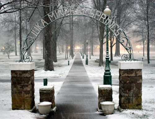 Snow-covered pathway leading through an archway in a park, surrounded by trees and soft lighting.