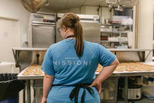 A person in a blue shirt with "we're on a MISSION!" stands in a kitchen, facing a table of cookies.